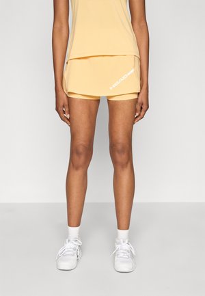 Shorts de sport jaunes à design superposé, avec un logo blanc sur le côté. Portés avec des baskets blanches et des chaussettes mi-mollet.