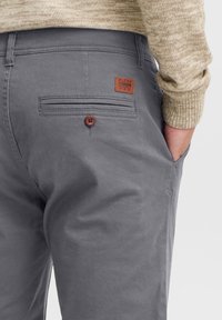 Pantalones de algodón grises con una etiqueta de parche de cuero marrón, detalle en el bolsillo trasero y botón marrón como acento. Textura suave y ajuste entallado.