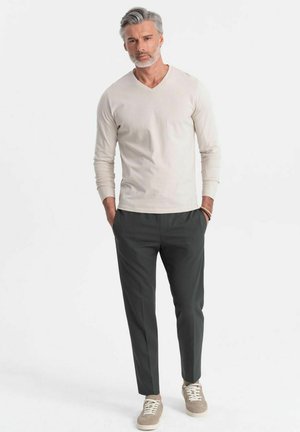 Camisa de manga larga beige claro con escote en V combinada con pantalones oscuros verdes a medida y zapatillas marrón claro con cordones blancos, de pie frente a un fondo blanco.