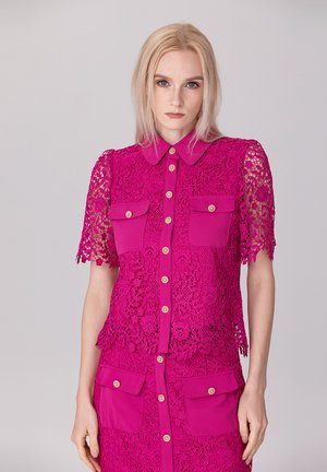Chemisier fuchsia en dentelle à manches courtes, design à boutons, deux poches avant et boutons dorés. Motifs floraux complexes sur l'ensemble.