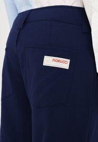 Temno modri hlače z gladko teksturo, z zadnjim žepom in belim etiketom, na katerem je v rdeči in modri barvi napis "Fiorucci Milano".