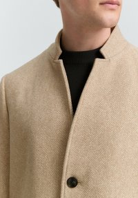 Beige wollen blend jas met een textuurpatroon, voorzien van een hoge kraag, een enkele knoopsluiting en een zwarte trui eronder.