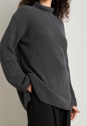 Personne portant un pull en maille côtelé gris foncé à col montant et un pantalon noir, main dans la poche, sur un fond uni.