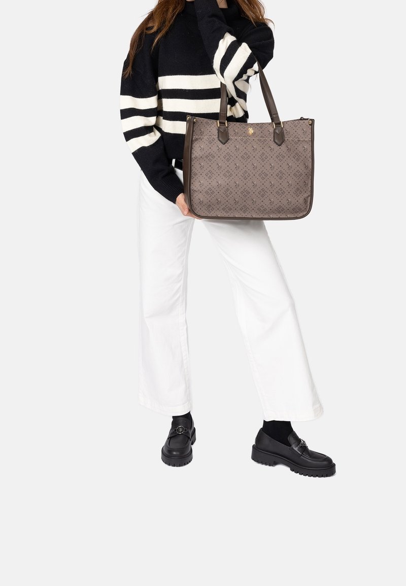 Femme portant un pull rayé noir et blanc, un pantalon blanc et des chaussures noires tient un grand sac cabas marron à motif avec des poignées foncées.