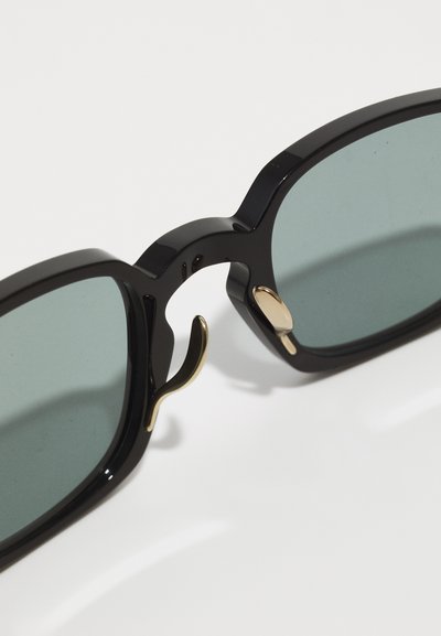 Schwarze Acetat-Sonnenbrillen mit grünen Gläsern, einer auffälligen rechteckigen Form und goldfarbenen Akzenten an den Rahmen und Scharnieren für einen Kontrast.