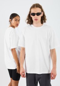 Bílé bavlněné trička s uvolněným střihem. Jeden model má na sobě černé šortky, druhý šedé kalhoty. Oba ukazují jednoduchý, čistý design.