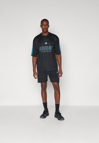 Melns Adidas sporta tērps, kas ietver īsām piedurknēm tīkla džersiju ar tirkīza akcentiem un logo, apvienots ar melniem šortiem un apaviem.