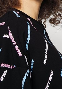 Persona con capelli ricci che indossa una maglietta nera con il testo multicolore "Karl Lagerfeld" stampato diagonalmente sul tessuto.