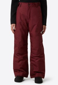 Pantaloni de zăpadă bordo, cu croială lejeră, confecționați din material rezistent la apă, prevăzuți cu închidere cu nasture și accente la cusăturile genunchilor. Sub pantaloni se văd cizme negre.