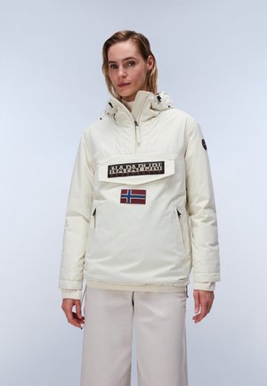 Woman wearing a cream-colored hooded Napapijri jacket with a front zipper pocket, logo patch, and Norwegian flag patch, paired with light pants.