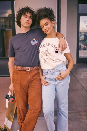 Két ember pózol együtt. Az egyik sötét pólót és barna kordbársony nadrágot visel, a másik krémszínű pólót fekete grafikával és világoskék farmert.