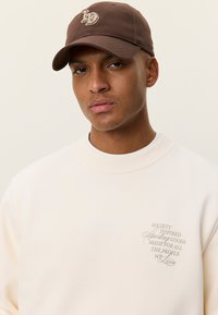 Μπεζ φούτερ με ριμπ λαιμόκοψη, με μπεζ κείμενο. Καφέ καπέλο με κεντημένο λογότυπο. Ομαλή υφή, άνετη εφαρμογή.