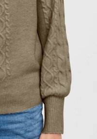 Olivgrüner Pullover mit Zopfmuster an den Ärmeln, gerippten Bündchen und Saum, kombiniert mit blauer Jeans.