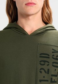 Oliivinvihreä huppari, jossa etutasku ja mustat painetut numerot. Pehmeä, sileä kangas, rento istuvuus ja kiristettävä huppu.