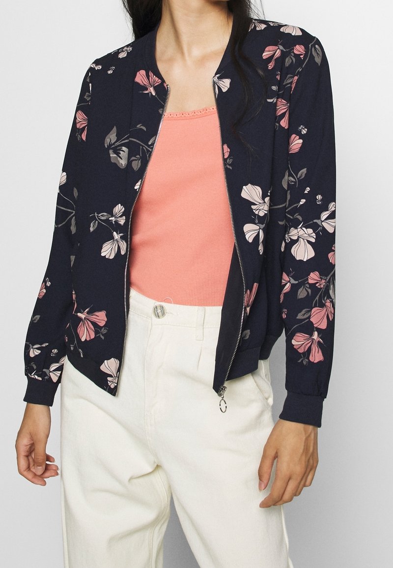 Marineblauwe bomberjack met bloemenprint in roze en beige. Heeft een ritssluiting aan de voorkant en geribbelde boorden, gedragen over een lichtroze top en witte broek.