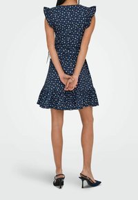 Marineblaues Kleid mit weißem Blumenmuster, gerafften Ärmeln, tailliertem Oberteil und ausgestellt Saum. Kombiniert mit schwarzen High-Heel-Sandalen.