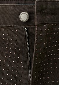 Donkere bruine broek met een ritssluiting en een rij decoratieve, glanzende studs in een patroon gerangschikt. Stevige stofstructuur zichtbaar.