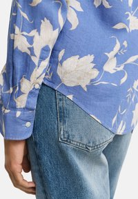 Blaues, blumig gemustertes Hemd aus leichtem Stoff, mit einem Knopfverschluss an den Manschetten und einem abgerundeten Saum, kombiniert mit hellblauen Jeans.