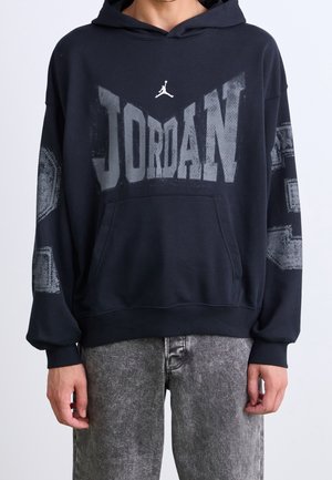 Person iført en sort Jordan hættetrøje med et stort falmet logo på brystet og gråvaskede jeans.
