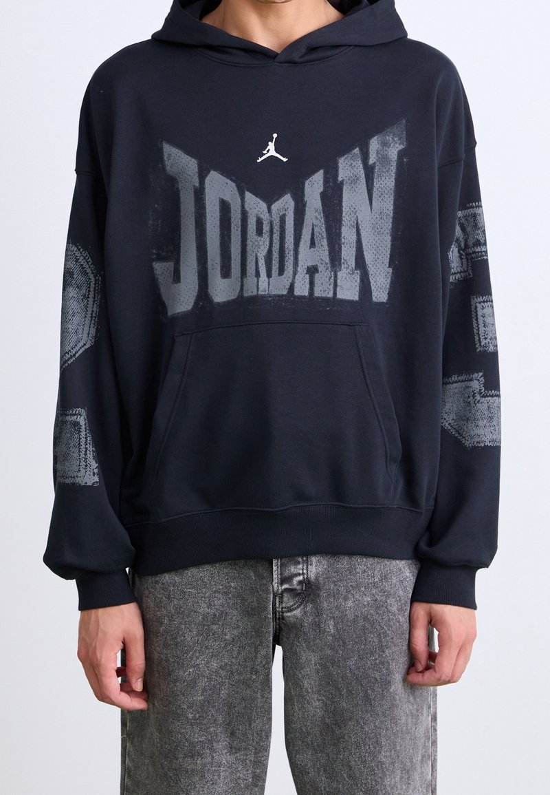 Personne portant un sweat à capuche Jordan noir avec un grand logo délavé sur la poitrine et un jean gris délavé.