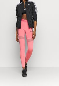 Chaqueta deportiva negra con rayas blancas, leggings rosa con paneles de malla y zapatillas negras con suela blanca, con el logo de Adidas.