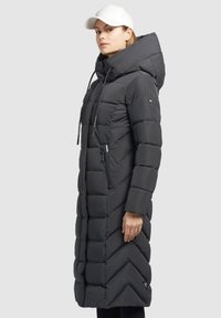 Lungo cappotto piumino nero con un motivo a chevron, dotato di cappuccio, lacci e una tasca con zip sul lato.
