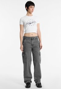 Iškirpta balta marškinė su "Guess Jeans" logotipu tamsiu atspaudų, kartu su pilkomis kargo kelnių, turinčiomis šonines kišenes ir laisvą prigludimą.
