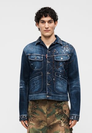 Νέος άνδρας με σγουρά μαλλιά φοράει ένα σκουρομπλε τζιν μπουφάν με καρφιά και παντελόνι παραλλαγής με μαύρα διακοσμητικά χάντρες.