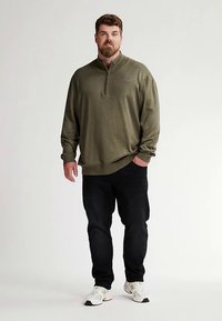 Oliivinvihreä vetoketjullinen collegepaita, jossa on resorit hihoissa ja helmassa, yhdistetty mustiin farkkuihin ja valkoisiin lenkkareihin, seisoo vaaleaa taustaa vasten.