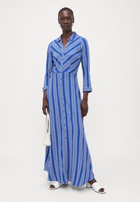 Blaues Maxikleid mit weißem Zickzackmuster, langen Ärmeln und einem Kragen. Gestylt mit weißen Sandalen und einer kleinen weißen Tasche.