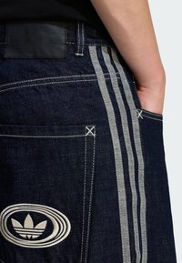 Jeans iz jeansa s temno modrim pranjem, kontrastnimi sivimi stranskimi črtami in vezenim belim logotipom na zadnjem žepu.