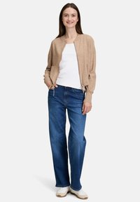 Frau steht lächelnd da, trägt einen beigen Reißverschluss-Pullover, ein weißes Hemd, blaue Jeans und weiße Turnschuhe vor einem schlichten hellen Hintergrund.