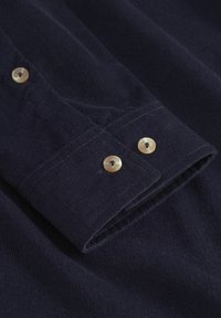 Tejido de corduroy en color azul marino con una superficie texturizada, que presenta dos botones de color claro en el puño y un detalle de manga doblada.