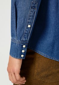 Manica di camicia in denim con polsino aderente e quattro bottoni a pressione bianchi. Tessuto blu con una texture classica e cuciture a contrasto.