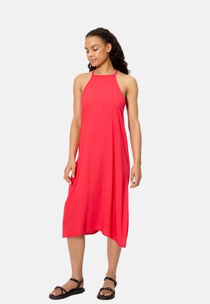 Mujer con cabello largo y rizado, vestida con un vestido midi rojo sin mangas y sandalias negras, de pie frente a un fondo liso y claro.