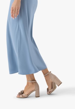 Stopy kobiety w beżowych sandałach na grubym obcasie z kwiatowym zdobieniem, ubranej w jasnoniebieskie spodnie z szerokimi nogawkami, stojącej na białym tle.