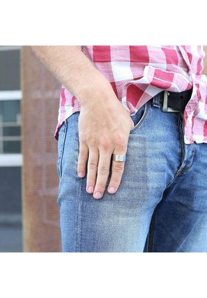 Zilveren ring op hand die rust op blauwe denim jeans, naast een geruit shirt in rood en wit. Stoftextuur zichtbaar op de kleding.