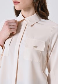 Blusa beige chiaro realizzata in un tessuto liscio e lucido. Presenta un colletto, due tasche applicate con dettagli di bottoni argentati e chiusura con bottoni sul davanti.