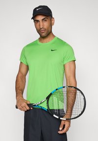 Πράσινη αθλητική μπλούζα Nike με κοντά μανίκια, με μαύρο λογότυπο. Το μοντέλο κρατάει μία ρακέτα τένις με μπλε λεπτομέρεια και φοράει ένα μαύρο καπέλο.