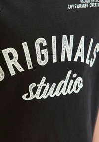 Camiseta de algodón negra con un gran texto blanco que dice "ORIGINALS" en una fuente en negrita y "studio" en un estilo cursivo, con una impresión texturizada.