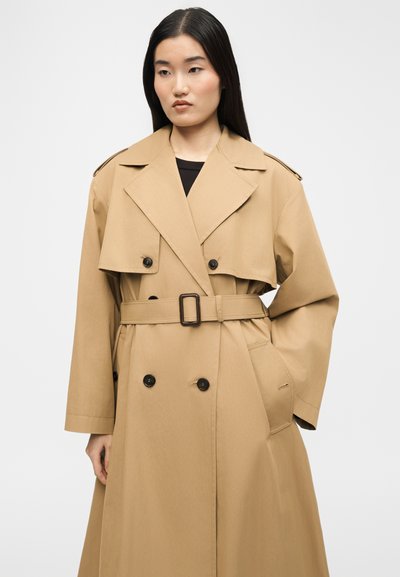 Γυναίκα με μακριά μαύρα μαλλιά που φοράει μπεζ καμπαρντίνα με ζώνη, μαύρα κουμπιά και φαρδιές πέτρες, στέκεται μπροστά σε ένα απλό φόντο.