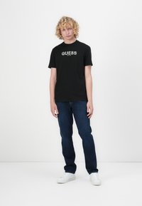 Juodas medvilninis marškinėliai su baltu "GUESS" logotipu, trumpomis rankovėmis, derinami su tamsiai mėlynomis džinsomis ir baltais sportbačiais ant vienspalvio fono.