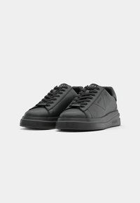 Paire de baskets basses noires à lacets avec semelles épaisses, affichant des logos discrets de la marque sur la languette, le côté et la semelle.
