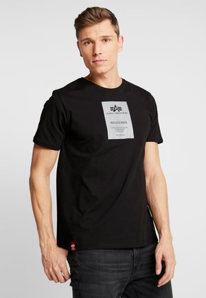 Schwarzes Baumwoll-T-Shirt mit Rundhalsausschnitt, versehen mit einem silbernen rechteckigen Grafiketikett und einem roten Akzentanhänger am Saum.