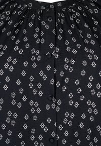 Chemise en tissu noir avec des motifs en forme de diamant blancs, dotée d'un col froncé et de cinq boutons noirs sur le devant.