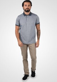 Grå piké med marineblå krage og ermer. Kombinert med beige bukser og sorte joggesko. Enkel, tettsittende design med en subtil logo.