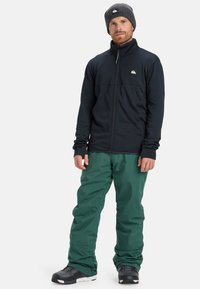 Schwarze Fleecejacke mit Reißverschluss und grüne wasserdichte Hose, kombiniert mit einer dunklen Mütze und schwarzen Schneestiefeln, geeignet für Aktivitäten bei kaltem Wetter.