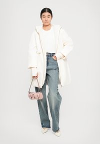 Witte oversized jas met capuchon, gelaagd over een wit shirt en blauwe high-waisted jeans. Heeft een gestructureerde roze tas en puntige witte hakken.