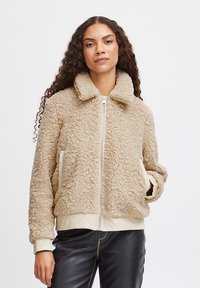 Smeđi faux shearling kaput s ovratnikom, prednjim zatvaračem, kremastim kviltanim rukavima i bočnim džepovima, nosi se preko crnih kožnih hlača.