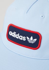 Casquette bleu clair avec une patch rectangulaire marine comportant le texte "adidas" en blanc et une bordure rouge, incluant le logo avec trois bandes.
